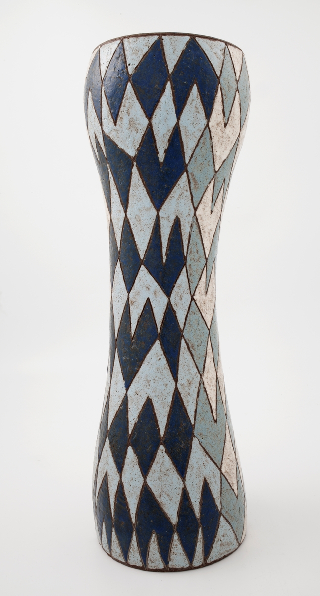 Høy, skulpturell vase i glasert steingods. Organisk sylinderform som slankes inn på midten, før den bli videst ved munning. Geometrisk, men uregelmessig harlequinmønster i mørkblått, lysblått og hvitt med mørkbrunt omriss (godsets bunnfarge) rundt hver fargeflate. Det brune skinner også delvis gjennom glasuren. Klar blå glasur innvendig.