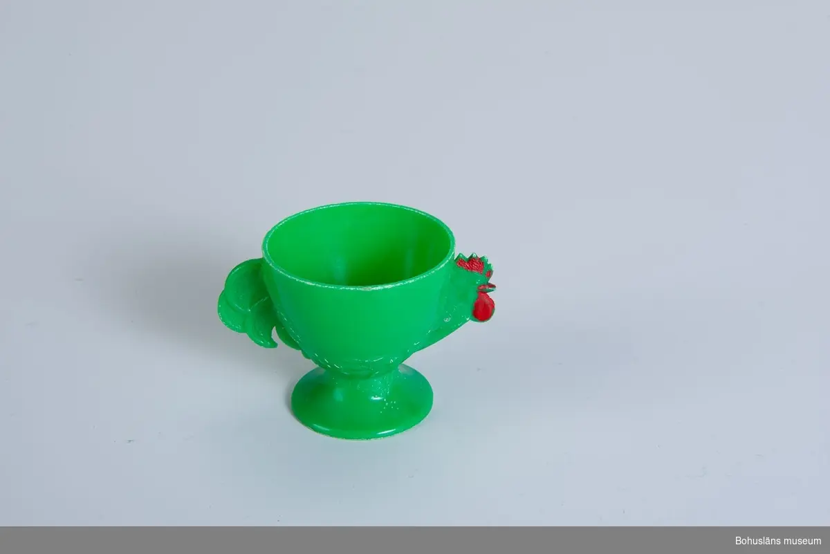 Äggkopp i i form av en tupp i grön plast med rödmålad näbb och tuppkam.
Märkning undertill: 
”Får en kokas en HELI-produkt 
Diskas i ljumt vatten”.