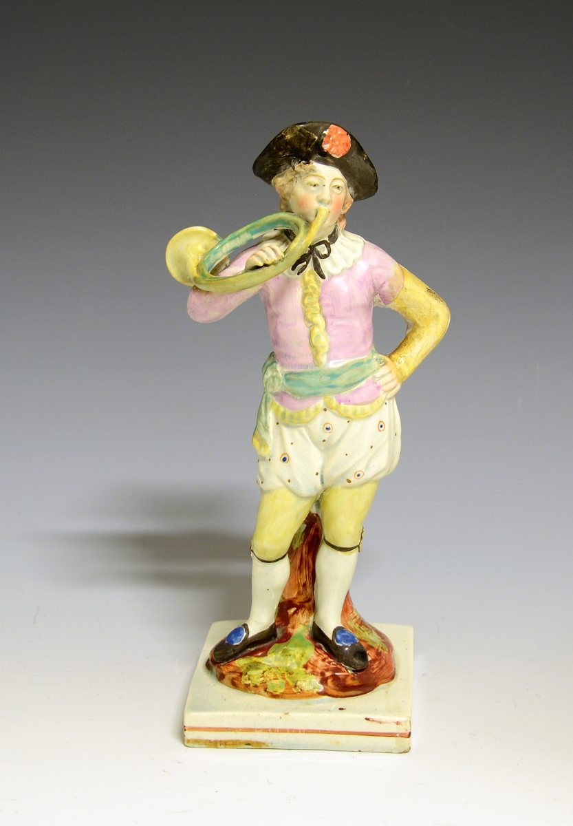 Mannsfigur av fajanse, blåser i et valdhorn som han holder i høyre hånd. Står på en kvadratisk sokkel.