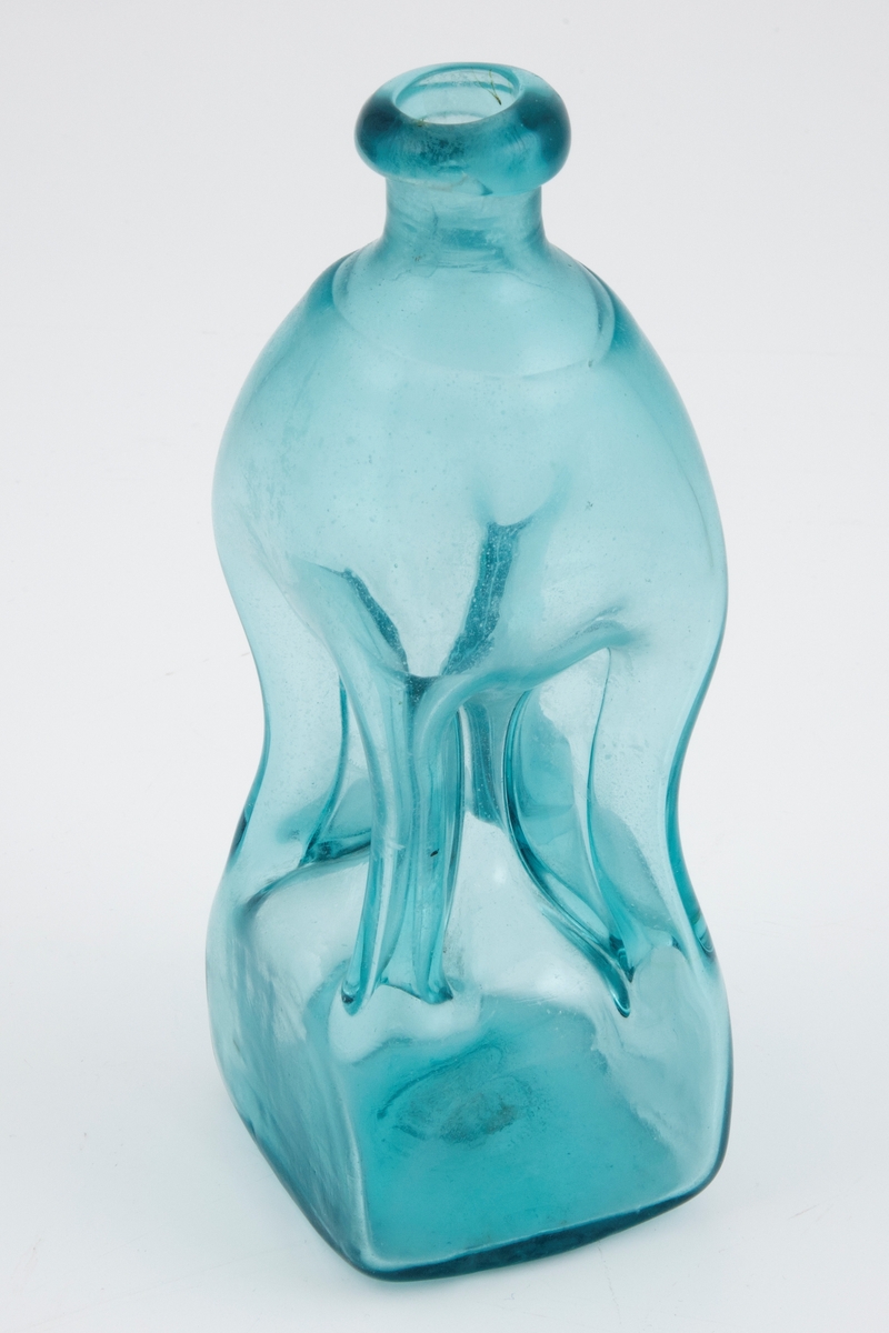 Klukkflaske i gjennomskinnelig lyseblått glass. Timeglass-lignende utforming, hvor den tilnærmet kvadratiske underdelen er forbundet med overdelen gjennom et tynt rør. Lav hals med glatt kantring, samt puntemerke på undersiden.