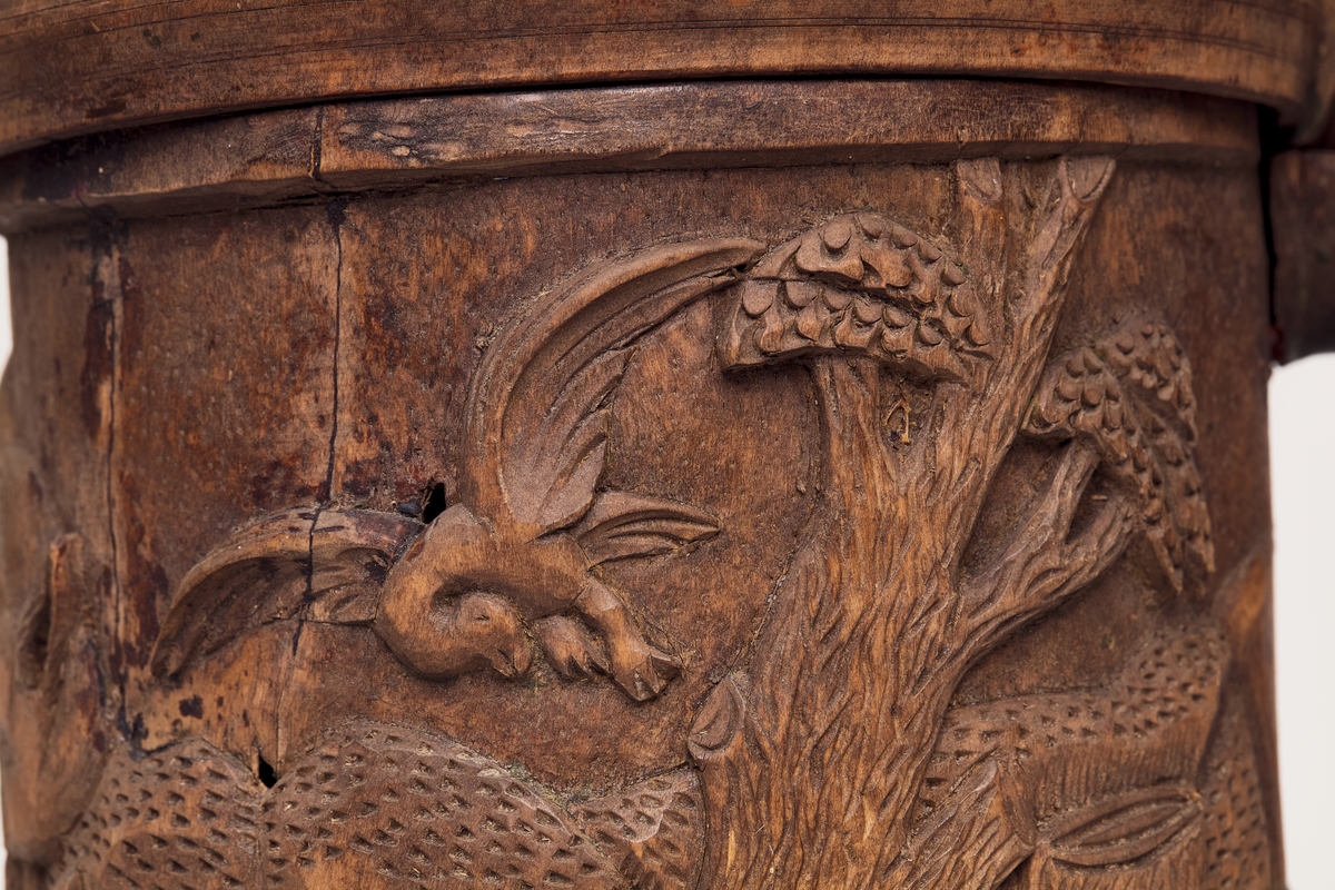 Kruset er sylinderformet og hviler på tre løver. Utskåret landskap med rev, fugl og trær, løveformet lokkgrep på lokket.