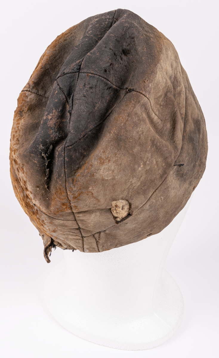 Mansmössa av skinn med rester av brun hårbeklädnad. Mössan är sydd av kilar (med många inkilade skarvar) och fodrad med vadderat, svart bomullstyg fastsytt på pappstomme. Mössan är tillplattad och något trasig. Den ger ett ålderdomligt intryck - tidigt 1800-tal.