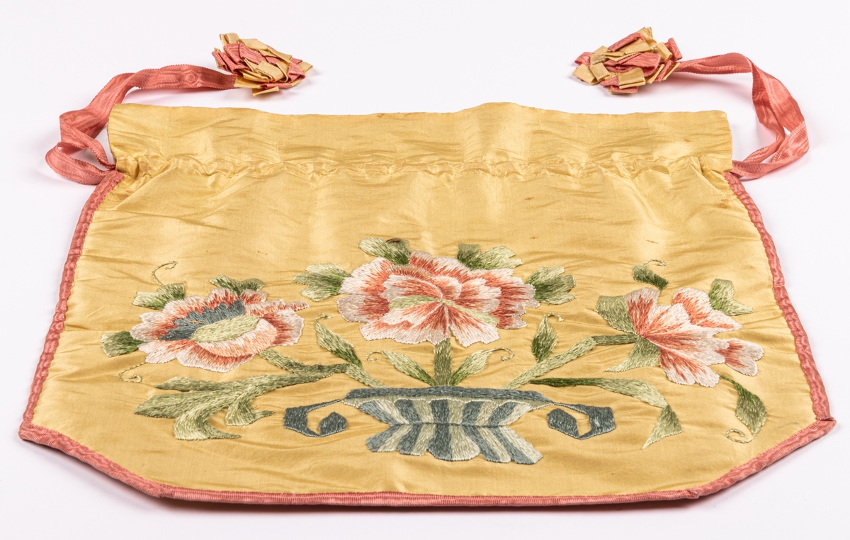 Väska av gult siden, broderad i schattersöm kantad med rosa ripsband som även utgör dragband och prydda med kokarder i rosa och gult, bodrad med grönt siden. Dragsko. 1890-tal.