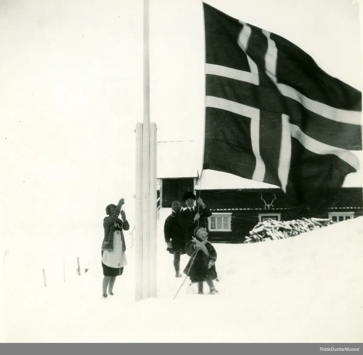 Njeallje olbmo norgga leavgga geassimen leavgastolpui.
Tre personer og en unge heiser opp det Norske flag.