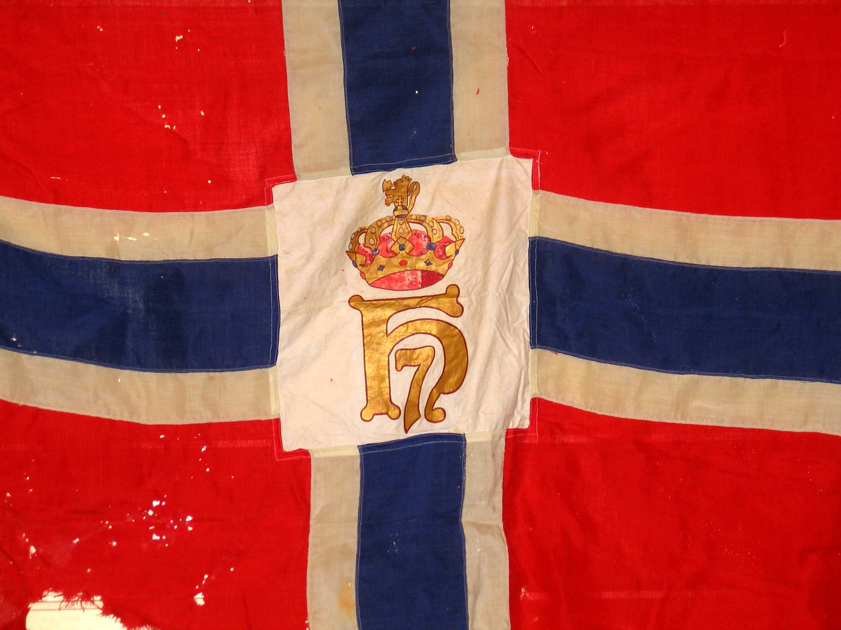 Norsk splittflagg med påsydd hvitt felt i midten. Motivet er trykt med silkestrykk i gull, rødt og blått. Motivet vises speilvendt på andre siden. Løpegang med snor og metallring.