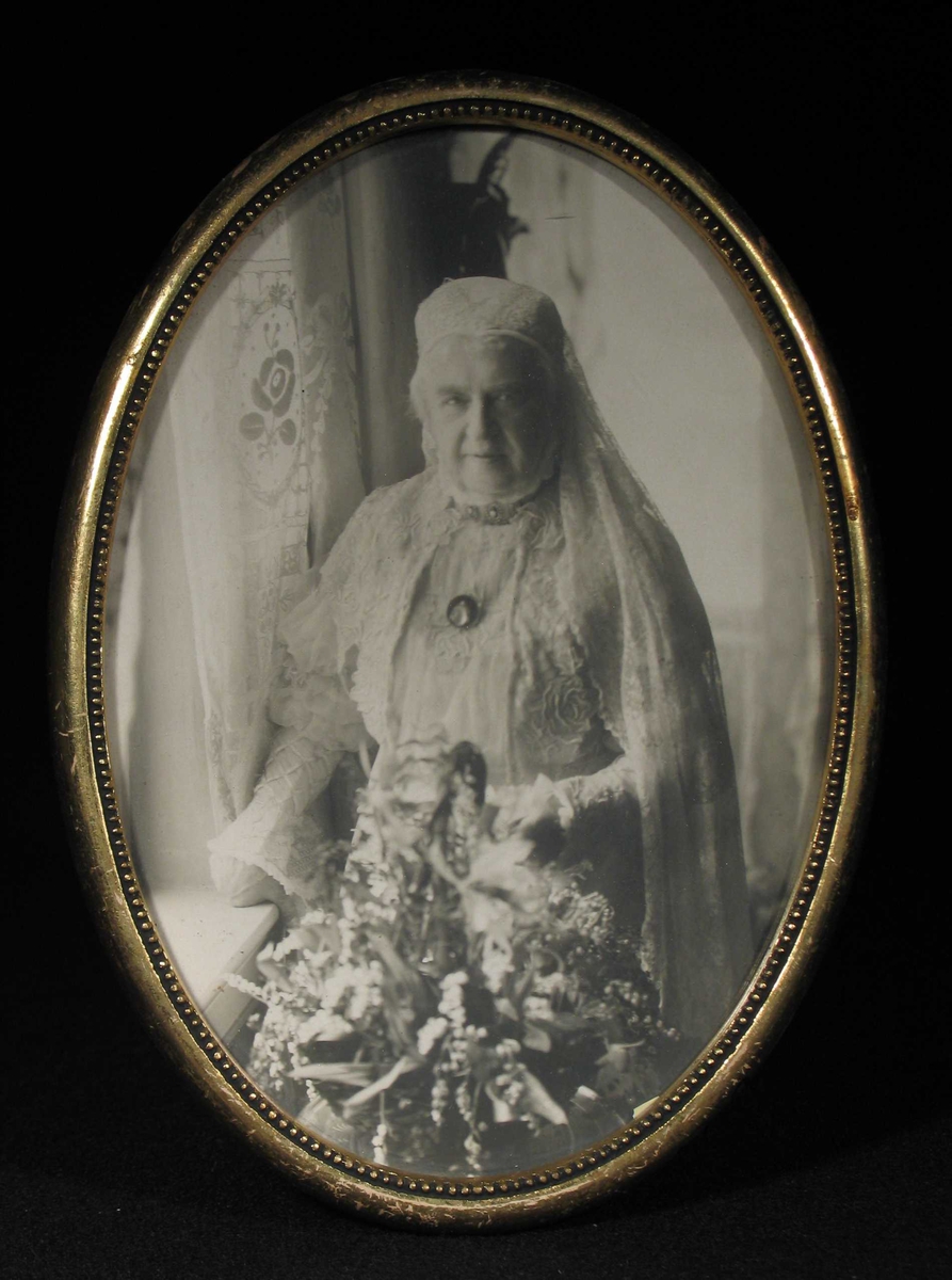 Eldre kvinne i hvit lang kjole stående ved et vindu med blomster i hånden.