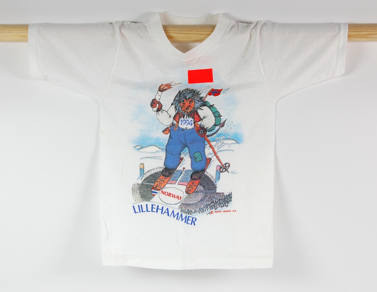 Hvit t-skjorte i barnestørrelse med flerfarget logo for de olympiske vinterleker på Lillehammer i 1994. Logoen domineres av motivet av et troll som tilsynelatende svever på ski over Lysgårdsbakken på Lillehammer. Det er klistret på en oransje prislapp med innskriften: 89.00.