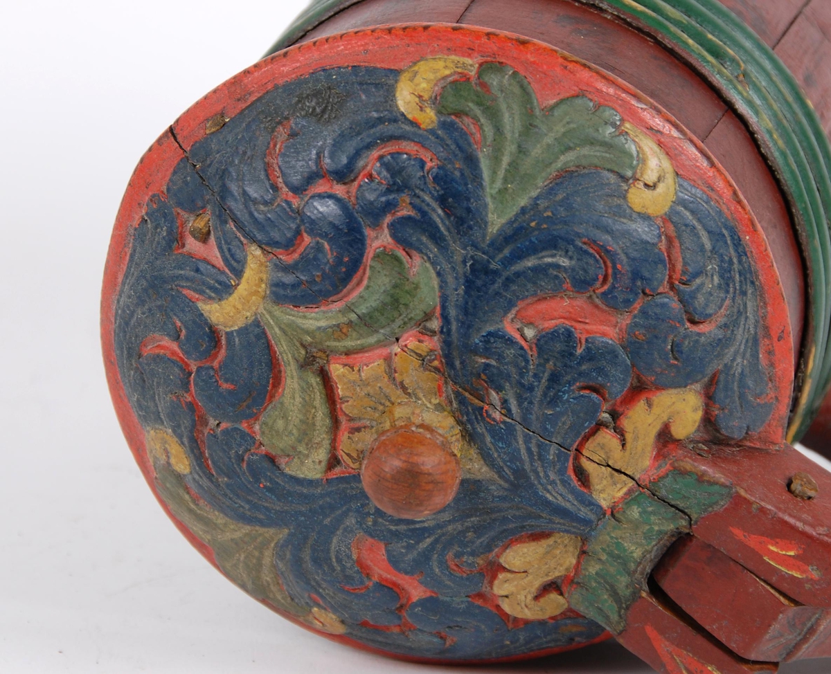 Kanne av furustaver. Lokk med akantusskurd malt i grønne, gule og blåe farger. Kuleformet lokkgrep. Korpus med fire grønnmalte gjorder oppe og en nede (opprinnelig tre). Hanken er utskåret av en av stavene, ender nederst i en plate med bladmotiver.