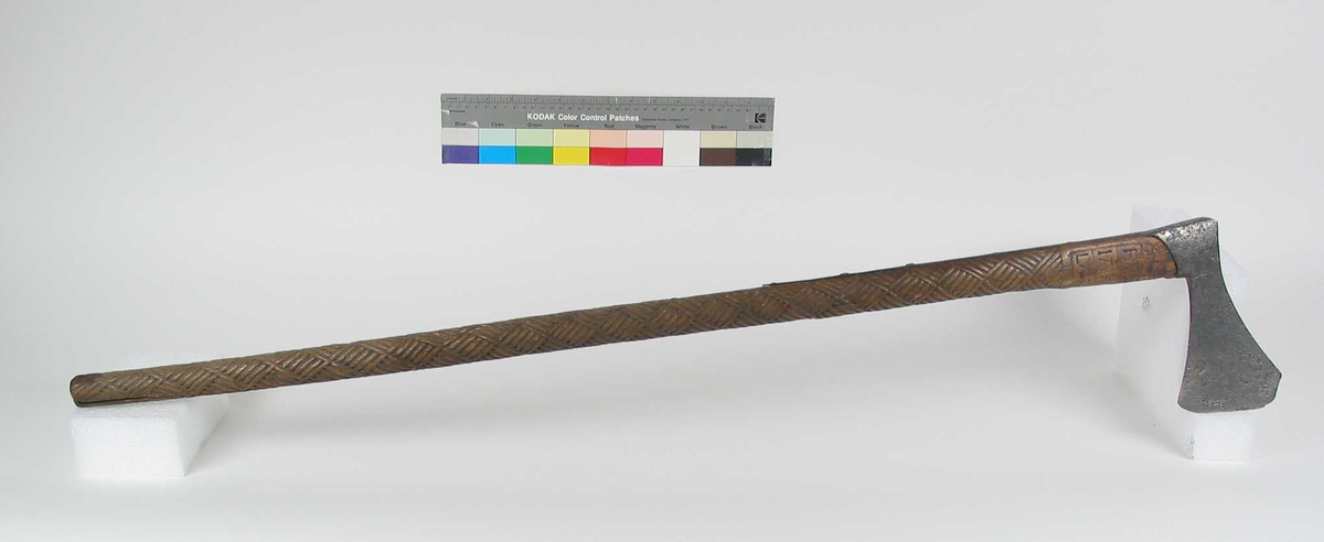 Asymmetrisk økseblad, sammensveiset med overliggende jernskinne.
Fremre øksebladspiss brukket.
Rikt dekorert skaft med flettet rutemotiv. Innskripsjon på øvre del av skaftet (se Påført tekst)
