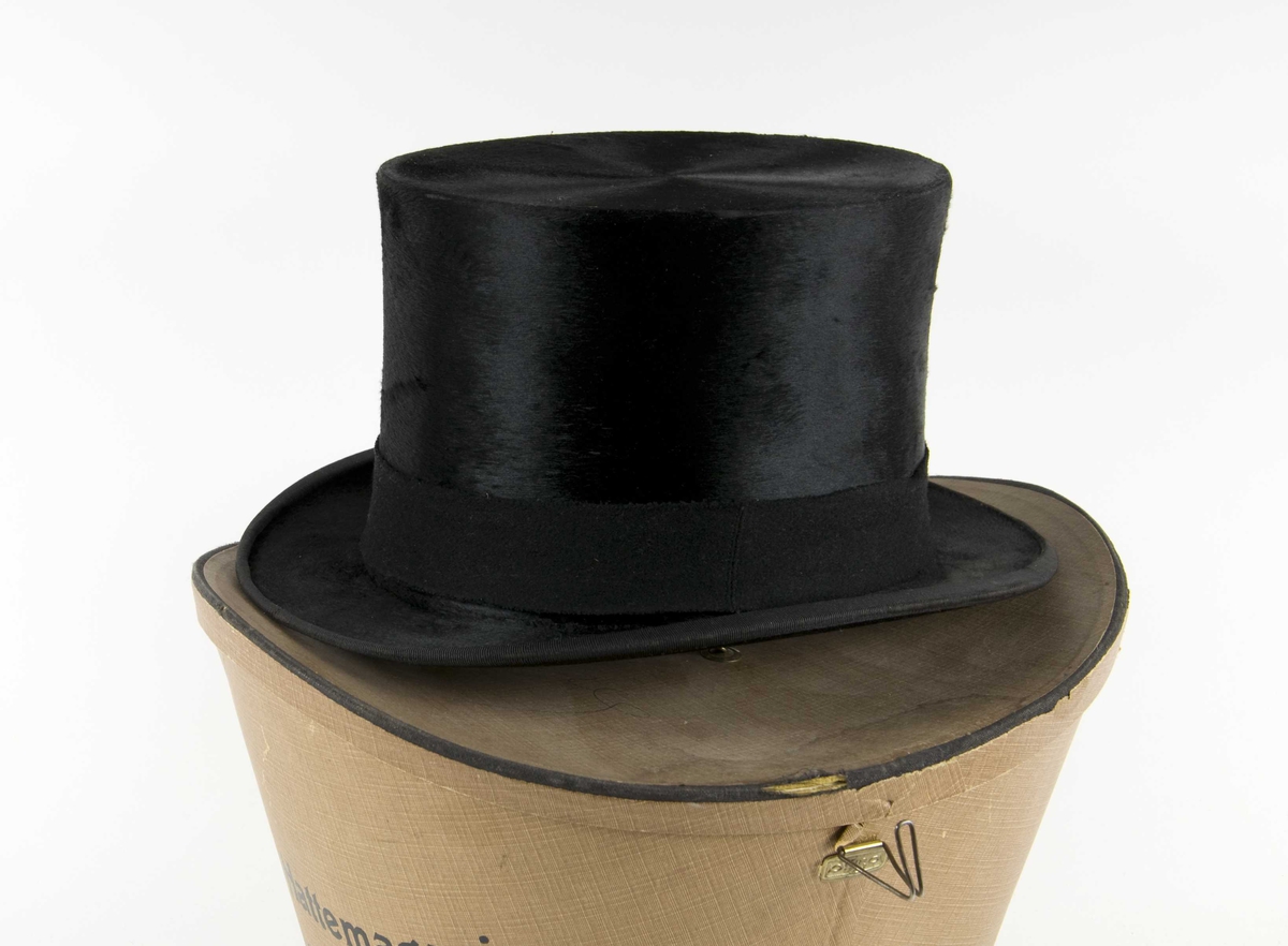 Sort flosshatt i hatteeske. Hatten har utvendig ullbånd. Hatten har svettereim av skinn og en sløyfe innvendig. En løs pute av grønn plysj i esken.