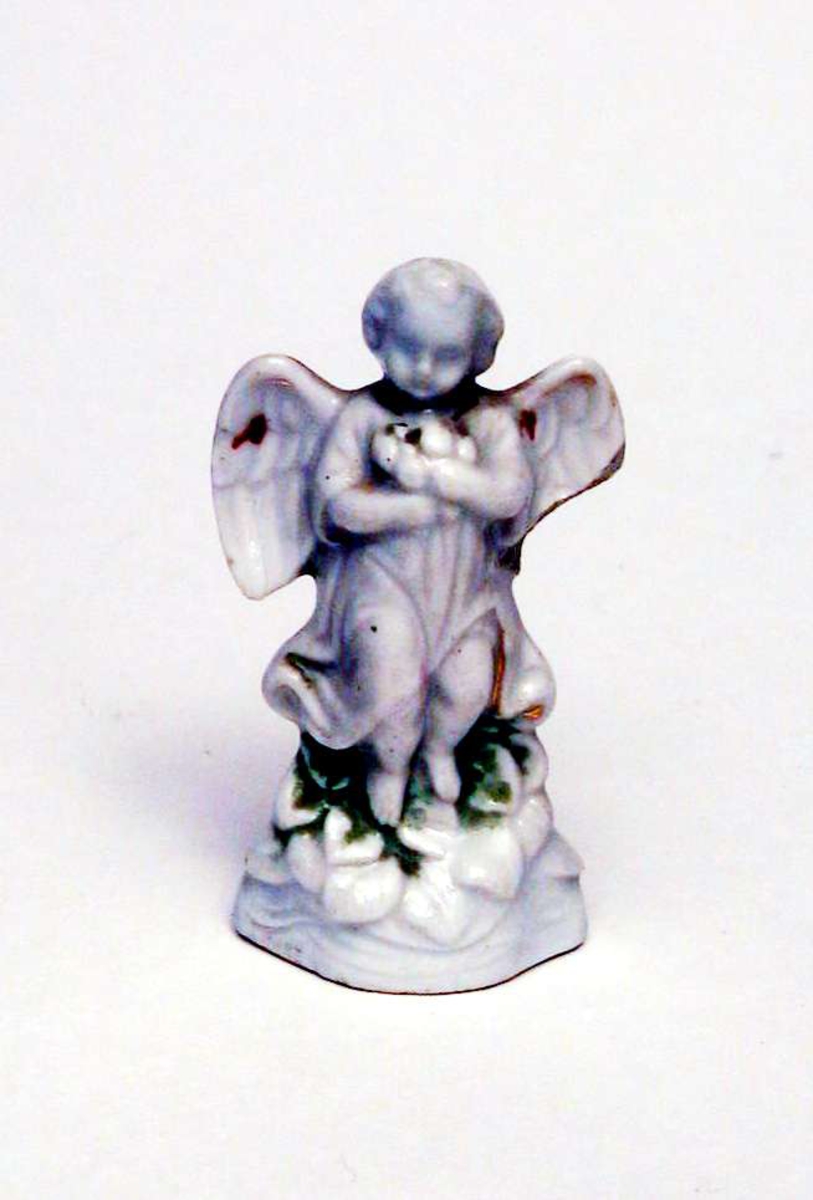 Liten porselensfigur, hovedsaklig hvit. Figuren forestiller en engel som holder en blomsterbukett. Deler av den ene vingen er slått av.