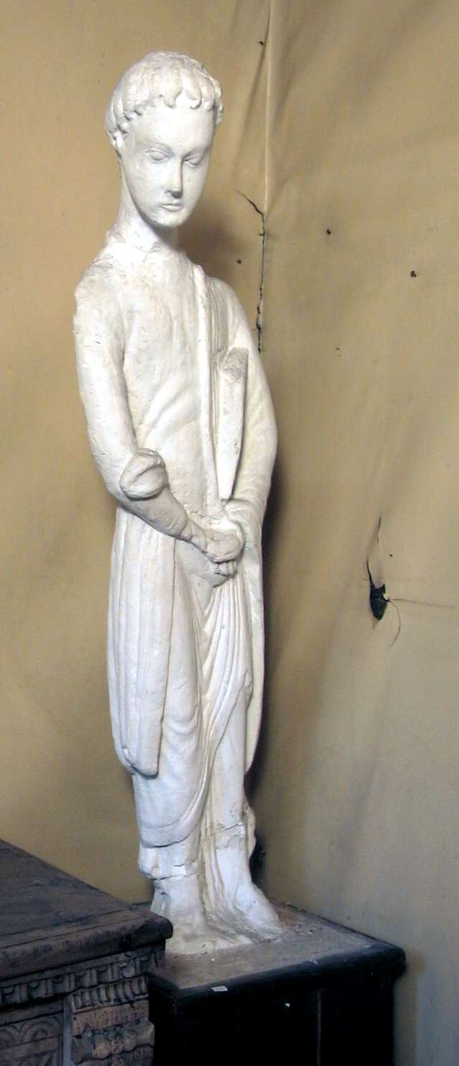 En avstøpning i gips av en treskulptur av evangelisten Johannes. Skulpturen står på en sokkel kledd med tre.