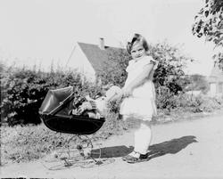 Liten pike med dokke i dokkevogn. Utendørs.