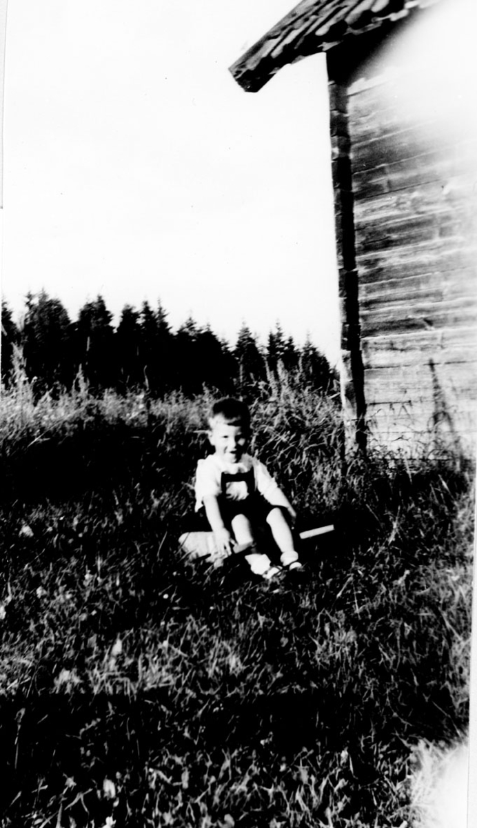 Husmannsplass, liten gutt sitter i gresset utenfor et hus.