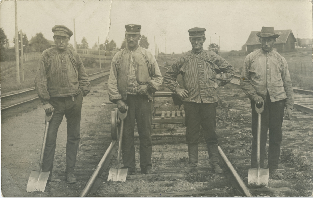 Fire menn med spader arbeider med jernbane spor. Dressin i bakgrunnen.