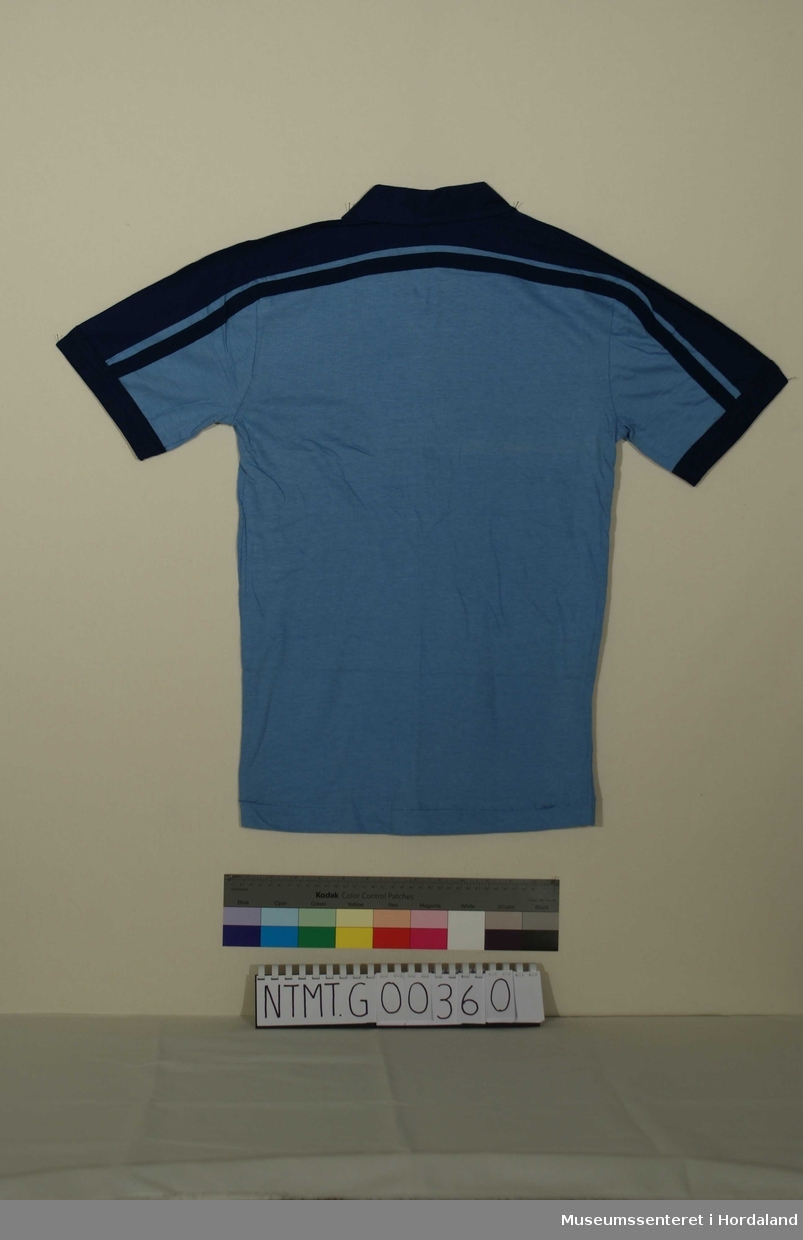 Lys blå t-skjorte med mørk blå krave, korte ermer, glidelåsåpning i hals, mørkeblått skulderstykke og kantavslutning.