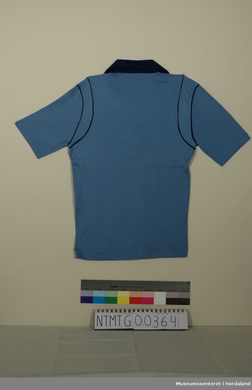 Lyseblå t-skjorte med mørkeblå krave, knappeåpning i hals med to knapper, mørkeblå pyntekanter ved halsåpning og ved ermefeste.