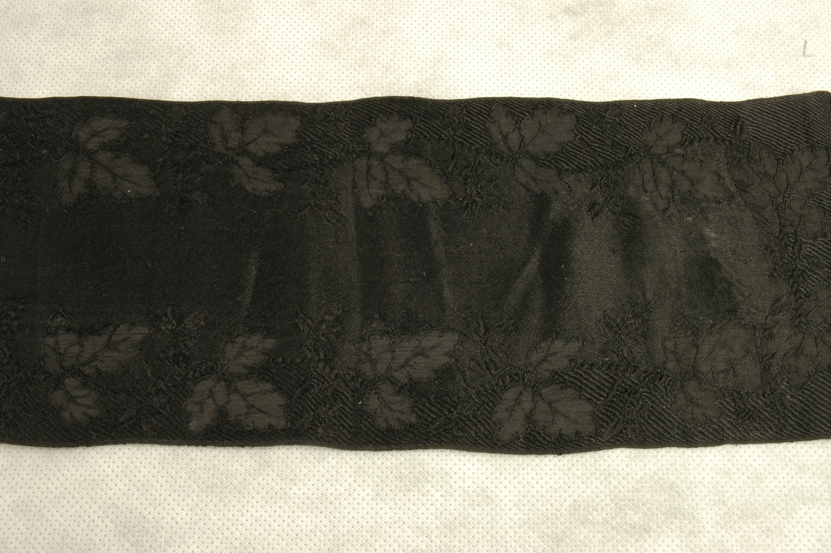 Svart mønstervevd silkeband med frynser. Bladmønster langs begge langsider.