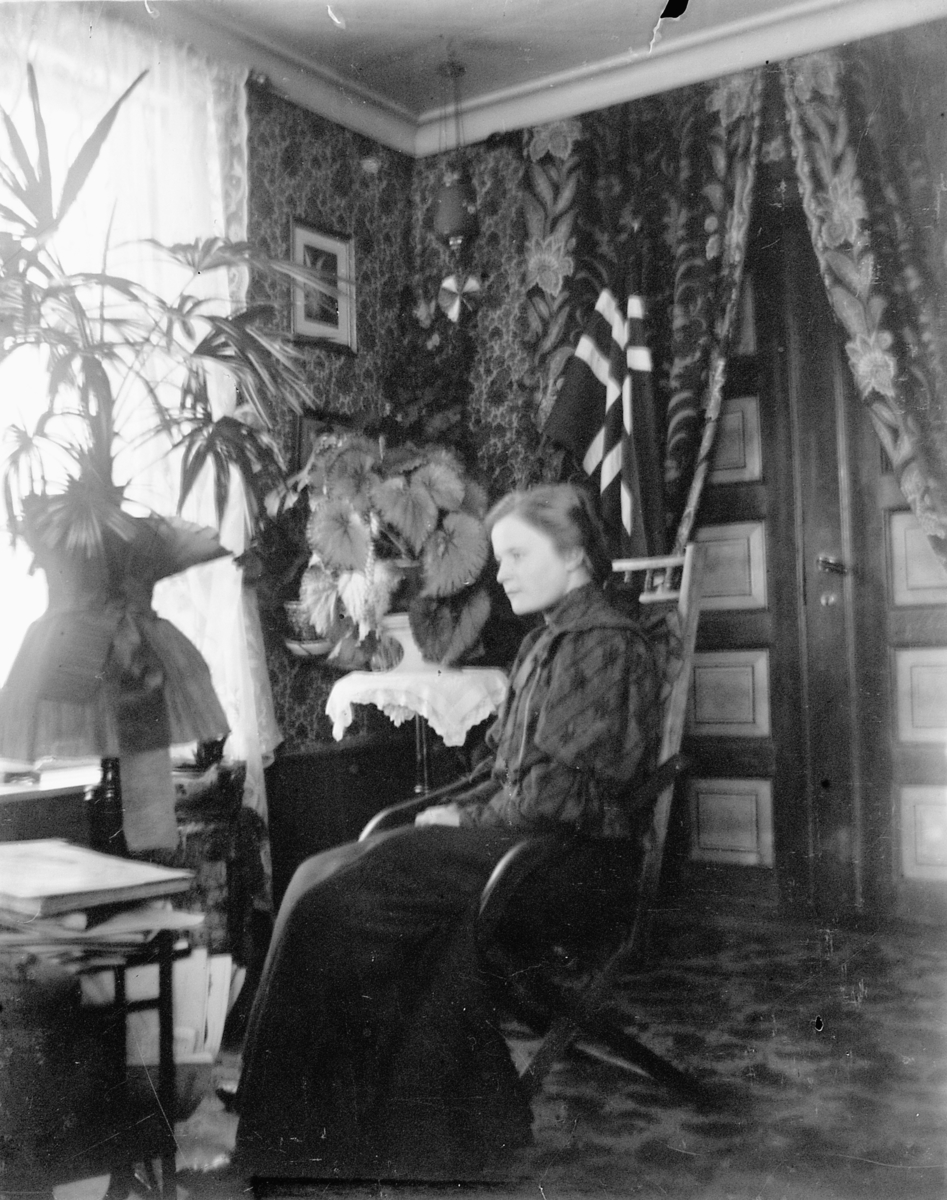 Interiør, stue. Anna Marie Johannessen, datter til bakemester Olaus og Lovise Johannessen, i gyngestol foran det norske flagget.