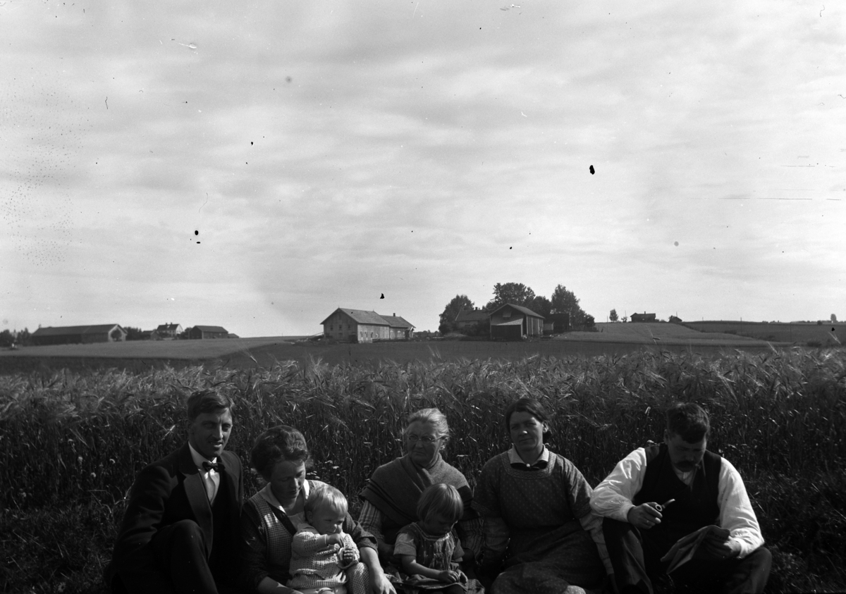 Ukjente personer sitter foran en kornåker. En gård i bakgrunnen. 