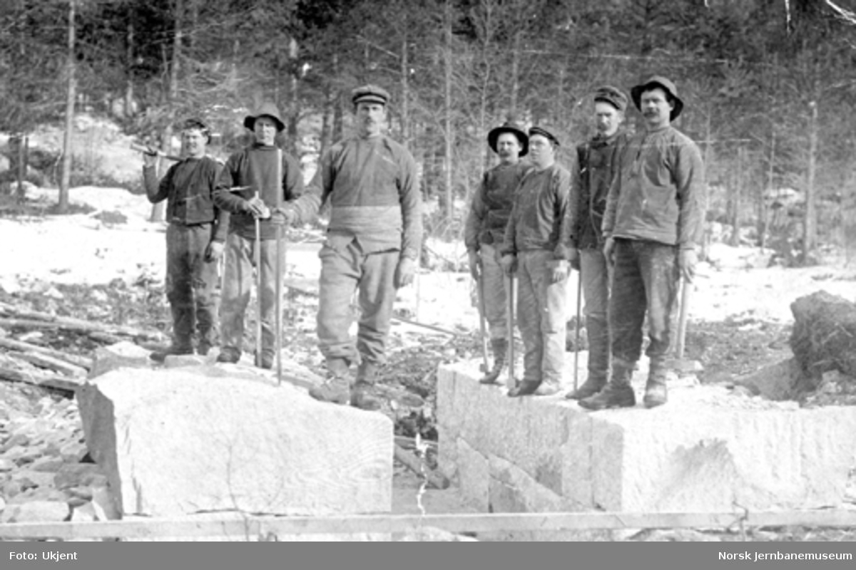 Sju anleggsarbeidere oppstilt for fotografering på steinblokker; trolig ved bygging av kulvert