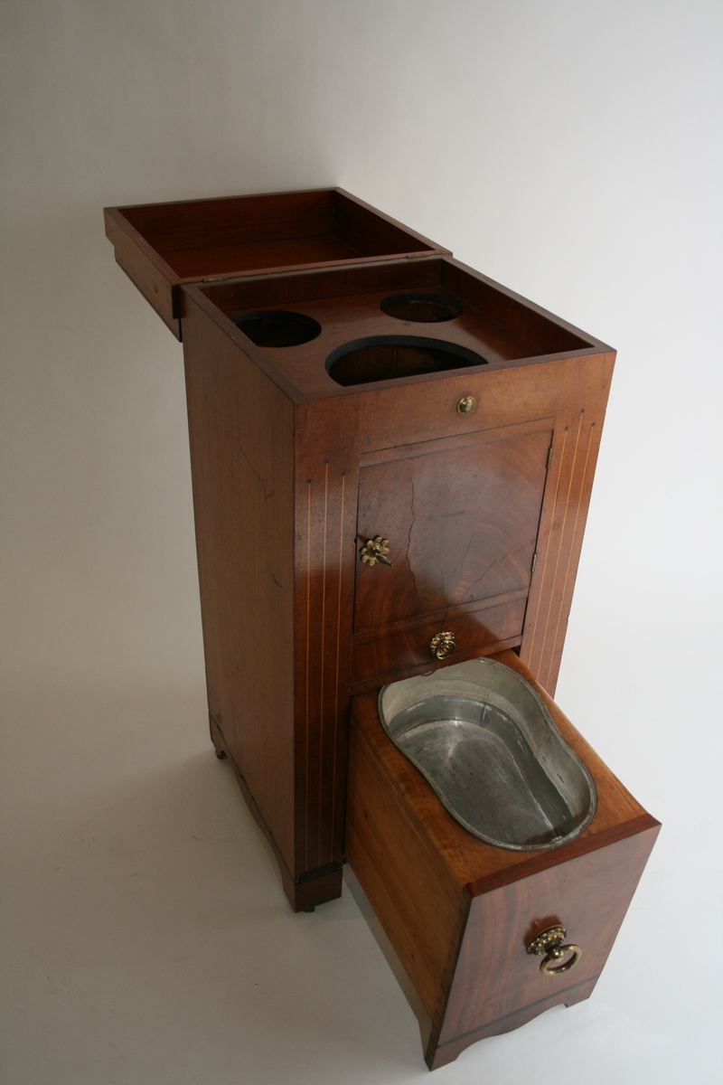 Toalettmøbel i mahogni. Toppplate til å åpne, skap og to skuffer. Nederste skuff har nattpotte eller bekken i blikk.
Forgylte bronsebeslag.