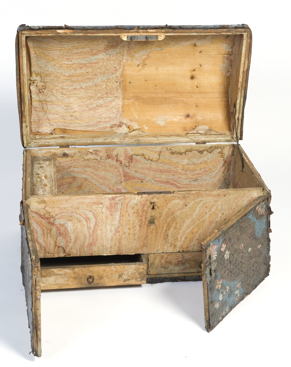 Kiste med rund lokk, lærtrekk, innside med marmorert papirtrekk.
Forside med to dører og skuffer.