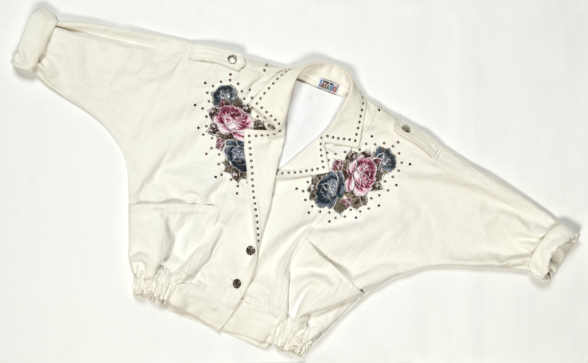Hvit jakke i bomull med dekor av kuler og fargestener med applikerte felt av blomstermønstret tekstil med glitter på. Tilhørende shorts med samme farger og dekor. 