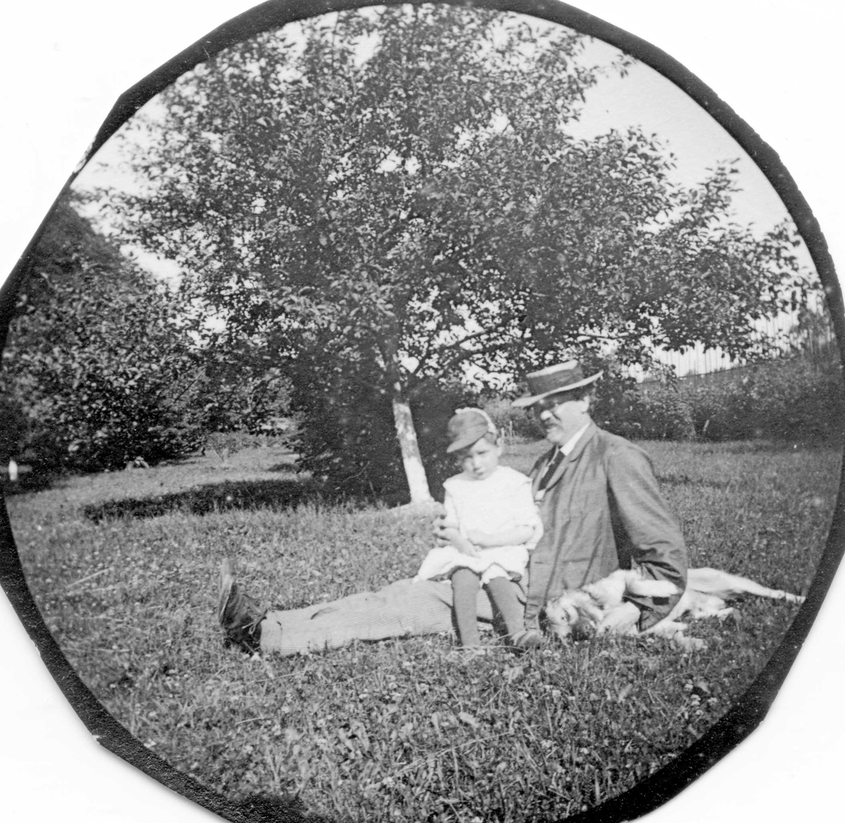 Dr. Elling Holst sitter i gresset med datteren Aagot på fanget, hund liggende ved siden av.