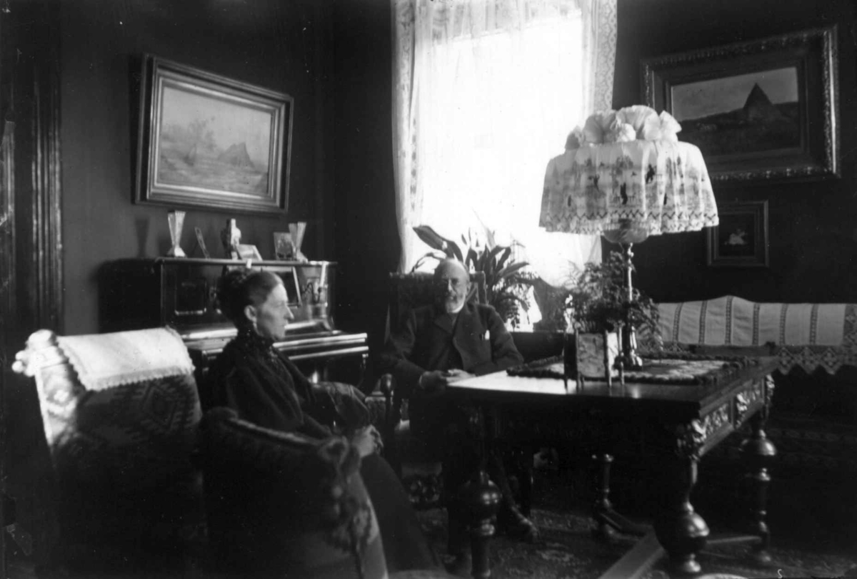 Interiør, ukjent sted. 1908-1910. Stue. Kvinne og mann ved bordet. Malerier. Piano.