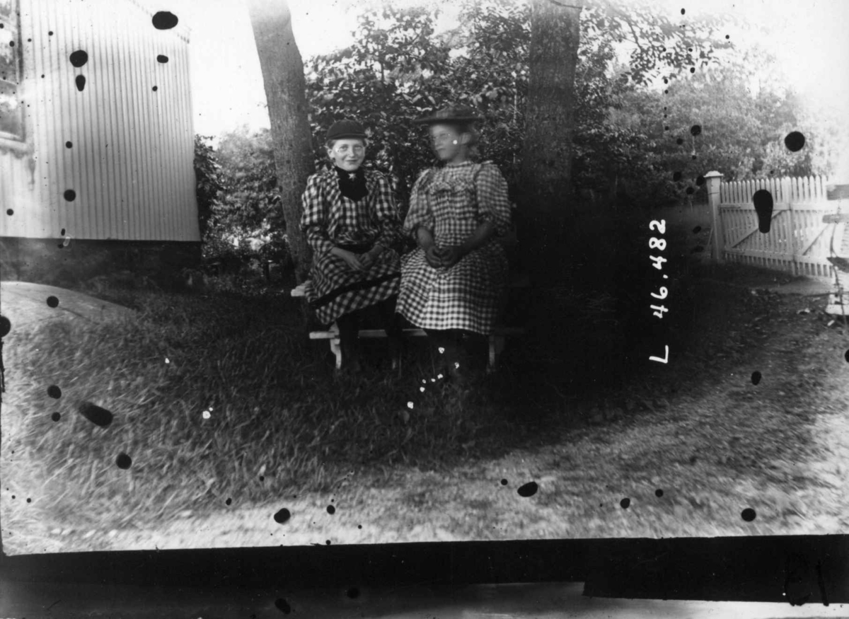 Drakt.1908-1910.To kvinner sittende i en hage. I kjoler og med hatter.
