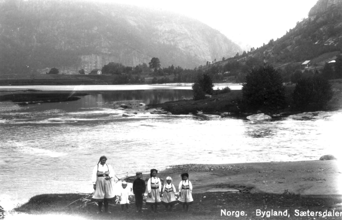 Gruppe i Setesdalsdrakter ved utløpet av Kvålsåni ca 1912 i Bygland. Til venstre Sigrid Neresdotter Bygland, sønnene Gunstein f- 1910 og Nere f. 1903, og døtrene Gyro, Birgit og Gunhild. Postkort