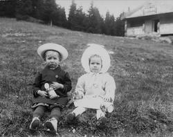 Barn sittende i helling, hus i bakgrunnen, ukjent sted.
Ser