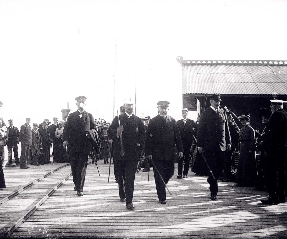 Fire menn i uniforn spaserer på brygga i Molde, Møre og Romsdal, foran tilskuere.