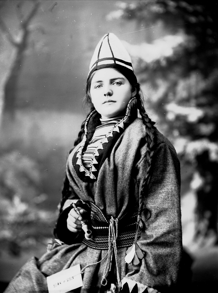 Kvinne i samisk inspirert drakt - kofte, lue og nålehus med beltering. Beltet er en beltestakk tilhørende en norsk bunad. I den ene hånden holder hun en pipe. Bakgrunnen er malt med trær.