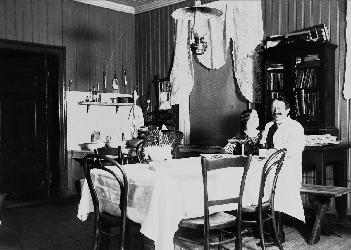 Interiør med mann og kvinne,  Axel Q. Wiborg med antatt sin kone Margrethe. Spisebord med vaskestell i bakgrunnen. Ukjent sted.