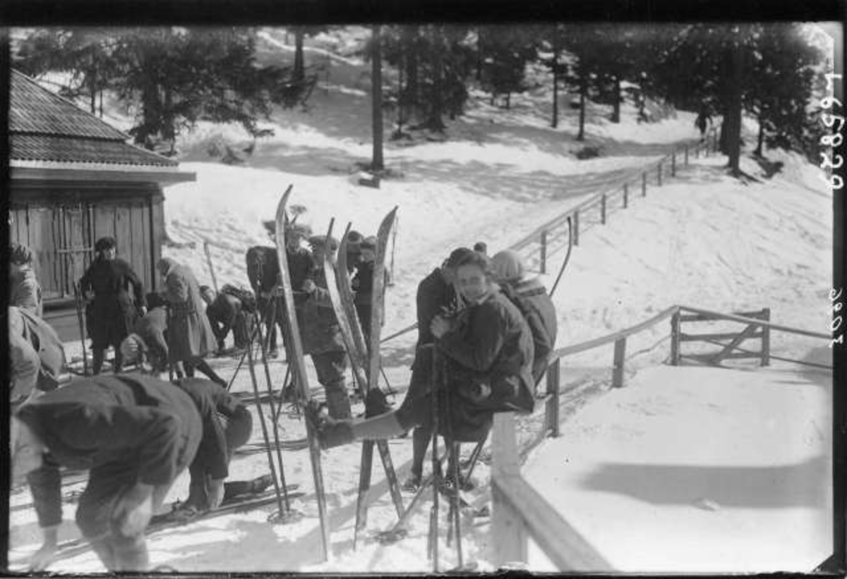 Skiløpere samlet utenfor skihytte i marka. Noen sitter på gjerdet, andre tar av eller på ski.