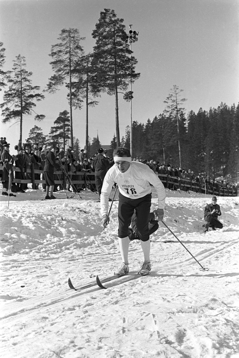 Skiløper med startnummer 76 i Holmenkollen. Publikum langs løypa.  Pressefotografer på sidelinjen. Holmenkollrennene 1963.