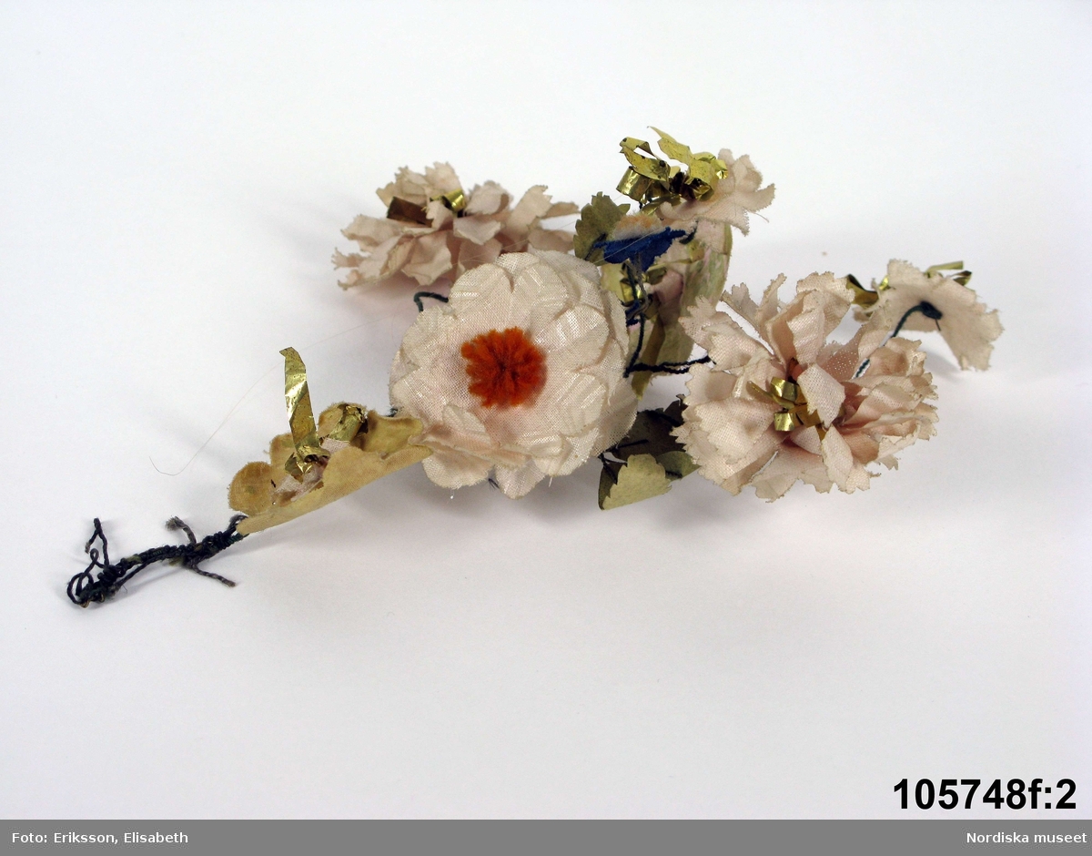 Hel bruddräkt bestående av 28 delar a-s.

a. Brudkrona,höjd 30 cm, diam 26 cm
stomme av ståltråd lindad med garn fäst vid en valk (b) klädd med svart yllekypert. Stommen helt klädd med konstgjorda blommor utstnsade i vaxat tyg med garntofsar som pistiller. De flesta blommorna är ljusa i vitt till rosa, omväxlande med blommor klippta av guldpapper.
c. mitt bak fastsydda långa lockar av cendréfärgat hår på ett löst band fäst på valken.
d. Trekantigt halskläde av vit tyll med maskinbroderi i 1820-30-talsstil med påsydd krage i samma form och stil. Runt kanterna men inte ända ut i snibbarna uddkant av två spetsar, den inre en maskinvävd spets, den yttre med runda uddar handknypplad av Vadstenatyp.
e. Bröstlapp, längd 37 cm, br. 17 cm, spets br. 10 cm
av ljust rosa glättat bomullslärft, ett rektangulärt stycke garnerat med guldspets som sytts fast i sicksack samt som kantning. I överkanten veckad tyllspets med trätt mönster.
f. 1-2. 2 lösa kvistar med samma typ av konstgjorda blommor som i kronan, och hör till denna.
g. Girland, längd ca 70 cm, ståltråd lindad med grön bomullstråd med påhängda konstgjorda ljusrosa blommor av vaxat bomullstyg med pistiller av röda och gula ullgarnstofsar med vita prickar på, blombladen av grönt papper.
h. Halsband längd 22 cm, pärlor diam, 8 mm. Röda träpärlor och vita glaspärlor uppträdda varannan på en bomullstråd som knyts runt halsen.
i. Halsband, längd 32 cm
vita halvtransparenta glaspärlor i fallande storlek med den största mitt fram, lås av mässing.
j. Örhänge, längd 3,5 cm, br. 2,5 cm
av avlånga pärlor, en vit tvärliggande och tre gula som hänger från den, två svarta prismaslipade ändpärlor samt flera små, små blå pärlor.
k. Halsremsa längd 58 cm, br. 4,5 cm.
smalt band av vit tyll med en fastsydd maskinvävd spets längs ena långsidan och en rynkad tyllremsa längs den andra.
l. Bälte, längd 81 cm, br. 4,7 cm.
band av ljust rosa glättat bomullslärft med två rader guldspets påsytt.
m.1. Rosetter, bandens br:  5 cm
2 ärmrosetter (nr 5 och 7) av röda sidenband i skiftande nyanser med invävda prickar. 2 armringar + en midjerosett med långa ändar samt en mindre rosett (på brudkronan) av ljusrosa sidentaft, blekta av utställning.
m.2. Rosett av rosa sidenband fäst mitt bak över lockarna på valken.
n. Näsduk, ca 42 x 28 cm. Tygbit av diagonalmönstrad tunn bomullsgas, rester av fåll i två hörn. Satt vikt, instoppad i ärmen på bruden.
o. Solfjäder
p. 1-2. Armkrås, ett par, br 10 cm.
1 par lösa ärmringar av tyll med trätt mönster, stärkta. Veckades och fästes i klänningsärmen.
q. 1-2. Ärmgarneringar, ett par, längd 22 cm, br. 25 cm. Ett smalt mittstycke av vit tyll med påsydda volanger åt båda håll av likadan tyll, kantade med smal maskinvävd spets.
r.Liv till brudklänning, längd fram 55 cm, mitt bak 40 cm, ärml. 45 cm. Av svart fint halvsidentyg, handsydd
2 framstycken, 2 bakstycken, isydda formsydda ärmar. Framstyckena med draperade djupa veck från axeln samlade i midjan till smalare veck som smalnar av i en snibb med isydda fjädrar som gör snibben styv. Sydd på foderliv av oblekt linneväv med styvnad i framkanterna. Fodret snört fram med 9 par mässingsmärlor dolda under knäppningen med hakar och hyskor.
Omfattande konsreveringslagningar på framstyckena. Har varit utställd sen åtminstone 1939 till 2004.
s. Kjol, längd 97 cm, vidd 280 cm, maskin-och handsömnad. Av svart mycket tunt ylle, ej detsamma som i livet vilket verkar äldre. Kjolen har 2 våder med vidden samlad bak i knäppta rynkor, slät framvåd, sprund i sidan, smal midjelinning knäppt med svart hake och hyska.
I bilagan finns en längre beskrivning hur bruden skulle kläs.
Foto finns av dräktfigur iklädd dräkten.

Berit Eldvik juni 2005


