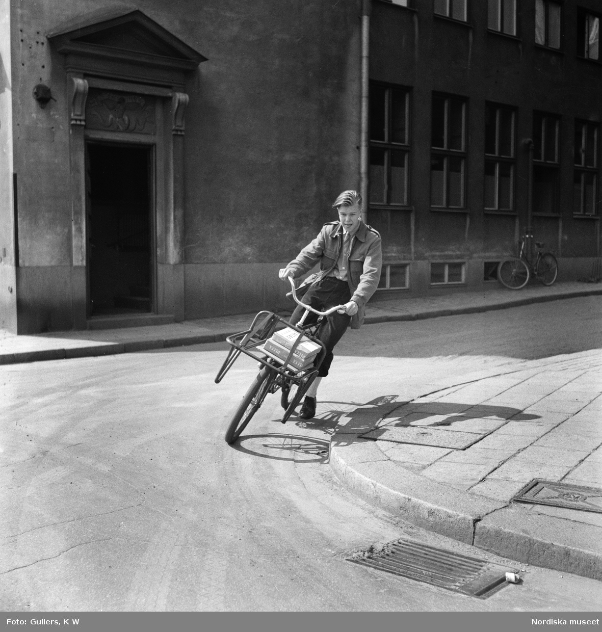Cykelbud i Stockholm. Bertil Parmryd 14 år svänger ut från Bredgränd i Gamla stan,  mot Skeppsbron i dåtidens vänstertrafik. På pakethållaren ligger paket med texten "Aktas".