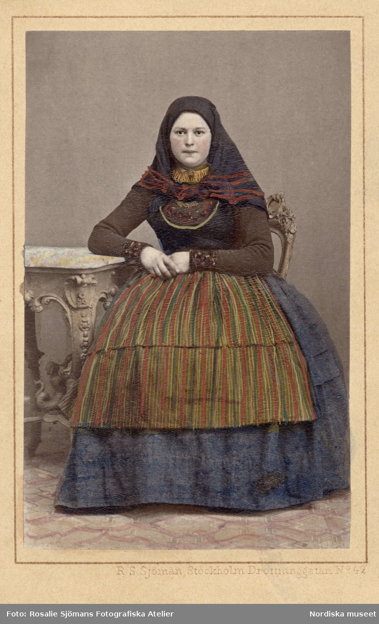 En kvinna i folkdräkt poserar sittande iklädd sjalett och randigt förkläde. Genarp, Skåne. Kolorerat i efterhand. Nordiska museet iv.nr. 18536C.