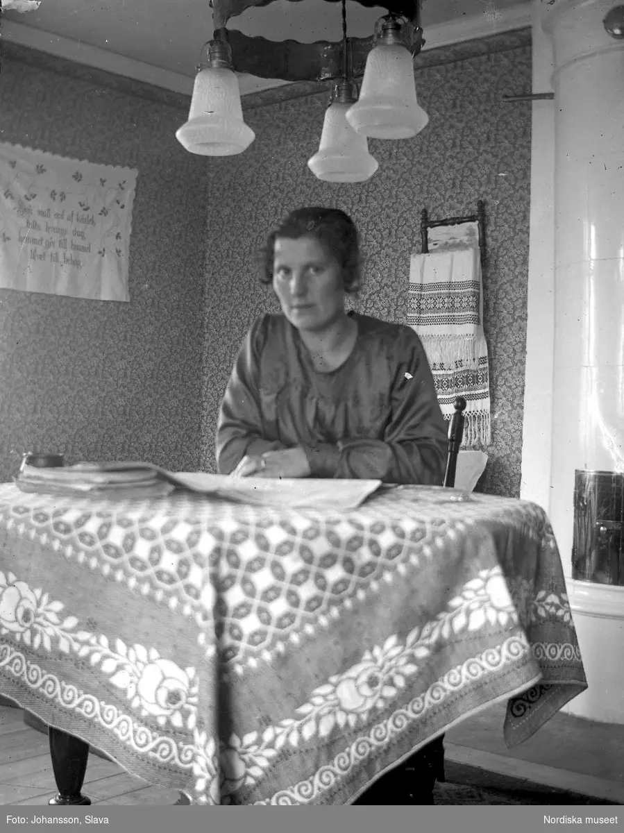 En kvinna sitter och läser en tidning vid ett bord. I bakgrunden mönstrade tapeter och en kakelugn. Lampa med tre glaskupor.
