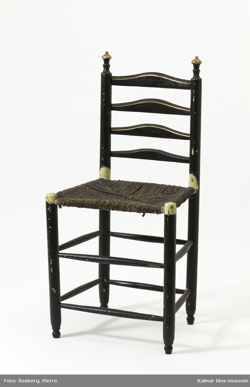 KLM 18989:1-2. Stol, stegstol, stegryggstol. Av typen Ladderback chair - stegryggstol. Två stycken. Av trä. Ryggstöd med fyra bladformade och tvärgående ryggbrickor. Ryggstolparna avslutas med snidade detaljer. Raka ben med dubbla rader av runtgående benslåar. Målad i svart med detaljer i guld. Under och intill sitsen är stolen målad i ljust gul, troligen originalfärg. Stol :1 har en flätad sits av rotting i tre lager. Stol :2 har en stoppad sits med undertyg av vävt ylle och övertyg av vinrött bomullstyg som är mycket skadat. Stoppad med halm.
