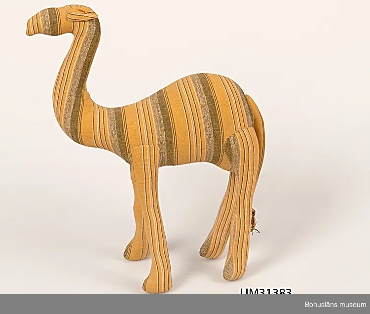 Kramdjur i form av långsmal kamel med svans. Sytt av gul- och brunrandigt bomullstyg, hårt stoppad med mjukt material, kanske kapock eller ull. Handsytt.