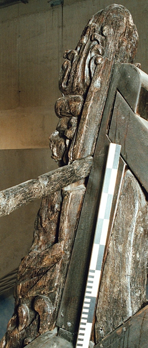 Skulptur i form av en hög, rikt snidad pelare eller stolpe, med kvadratisk genomskärning.

Tre av pelarens sidor är skulpterade medan den fjärde utgör anliggningsyta, försedd med ett antal olikformade urtag. De skulpterade sidorna är horisontellt indelade i tre eller fyra sektioner eller nivåer, avgränsade från varandra av profilerade och snedställda vulster. Nedtill avslutas skulpturen av en ornerad och på mitten insvängd formation.



Ovanför denna nedre avslutning är pelaren utformad som en reslig mansfigur, vänd mot vänster, sedd från betraktaren. Mansfiguren, som täcker tre av skulpturens fyra sidor, är iförd en romersk rustning med en bred fältbindel draperad från höger axel snett över bröstet. På huvudet med det lockiga och utslagna håret sitter en krona av öppen typ, med liljeliknande ornament. Figurens vänstra arm är böjd, medan den högra armen är sträckt utmed sidan. Handen till den högra armen saknas, men har hållit en rikt profilerad sköld, med mycket slitna och vaga konturer av en på bakbenen mot vänster gående grip med stora vingar. Skölden är snidad i ett med figuren, men med kraftiga underskärningar. Vid figurens fötter skymtar ett djur av något slag. Bakom mannens huvud tycks ett snidat textilstycke, kanske del till ett draperi, vara anbragt.



Ovanför den romerska krigaren eller riddaren har pelaren formen av en herm utan armar med långt, krusigt hår och mustaschprytt ansikte. Liksom mansfiguren täcker även hermen tre sidor av pelaren. Huvudet är vridet mot vänster. Överkroppen är bar så när som på ett flikigt plagg som täcker axlar och övre delen av bröstet. Från huvudet och runt midjan är ett tygstycke draperat. Hermens nederdel är formad till en stor maskaron med uppspärrad mun och utsträckt tunga. Maskaronen övergår upptill och utmed sidorna i yppiga bladformationer.



Pelaren avslutas upptill av ett knoppliknande ornament. Ornamentet är ornerat med akantusbladliknande bildningar samt fyra byglar som möts på toppen, där vissa spår antyder att det ursprungligen suttit ett klot eller dylikt, men som fallit bort. Eventuellt kan ornamentet utgöra en huvudbonad till hermen.

Skulpturen är kraftigt sliten.



Text in English: Sculpture in the shape of a richly carved column or pillar.

Three sides of the pillar are carved, while the fourth side comprises the attachment surface and has a number of variously-shaped indentations. The sculptured sides are divided horizontally into three or four sections separated by angled tori.



The lower section of the pillar is carved in the figure of a tall man. The figure is clad in Roman armour and has a field badge (cloak/plaid) draped across his chest at an angle from his right shoulder. On his head he wears an open crown. He has long, curly hair. The right arm is broken off, the left is at his side. The left hand is missing but once held a richly carved shield.

The shield is carved out of the same piece of wood as the figure, but deeply undercut. A carved figure is apparent on the shield. Although the shield is badly worn, it is possible to recognise one of the hind legs of a griffin with large wings. At the man's feet lies an animal of some kind.



Above the Roman warrior or knight, the pillar is carved in the shape of a herm, without arms and with long, curly hair and a moustache. Like the male figure, the herm also covers three sides of the pillar. The head is twisted to the left. The upper part of the body is bare except for a piece of cloth that covers the shoulders and upper chest. The lower part of the herm is carved as a large mask, its mouth open and tongue protruding. The sides and upper parts of the mask are carved as luxuriant leaves.



The upper part of the pillar has a ornament in shape of a knob. The ornament has a ring of acanthus leaves and four spherical loops run vertically from the acanthus leaves and meet at a point at the top. An object, for example a globe appears to have adorned the top of the ornament. This object is missing. The ornament may also be a part of the headgear of the herm.

The sculpture is severely worn.