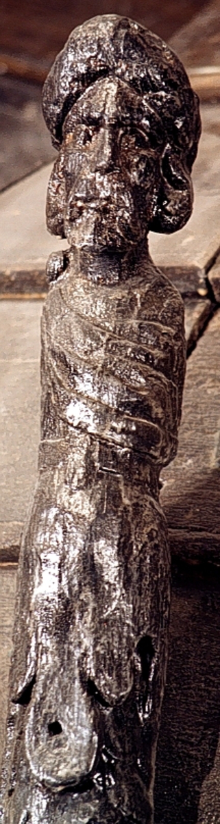 Skulpterad triton med turbanliknande lagerkrans på huvudet.

Huvudet har långt, ringlande hår samt stora och nedhängande mustascher. Snett över bröstet är en klädnad draperad. Klädnaden är fästad i en knut på höger axel. Kring livet sitter ett band med en oval.



Kroppens övergång till de dubbla, snodda fiskstjärtarna döljs av ett stort rundflikigt akantusblad. Huvudet är till största delen framställd som rundskulptur, dock något avplanad. Från nackhöjd och halvvägs ned på partiet med fiskstjärtarna är baksidan avplanad och slät - delvis något skålad - varefter det sitter ett snett urtag. Fotändan är vinkelformigt skålad vilket ger den en V-format tvärsektion, nedsänkningen är dock inte symmetriskt placerad. Nedtill böjer fotänden av inåt.

Skulpturen är mycket välbevarad.



Text in English: Sculpted Triton with a turban-like laurel wreath on its head.

The figure has a large pendulous moustache and long curly hair. There is a garment draped diagonally across the chest and fastened in a knot on the right shoulder. There is a girdle or belt round the waist with an oval centrepiece.



The transition between the body and the double fish tails is concealed by a large rounded acanthus leaf. The majority of the head is carved as a three-dimensional sculpture. The rear side surface has been planed smoothly.  The foot end has been hollowed out at an angle, giving it a V-shaped cross section, the hollowed-out section is not, however, symmetrically placed. The foot end curves inwards.

The sculpture is very well preserved.
