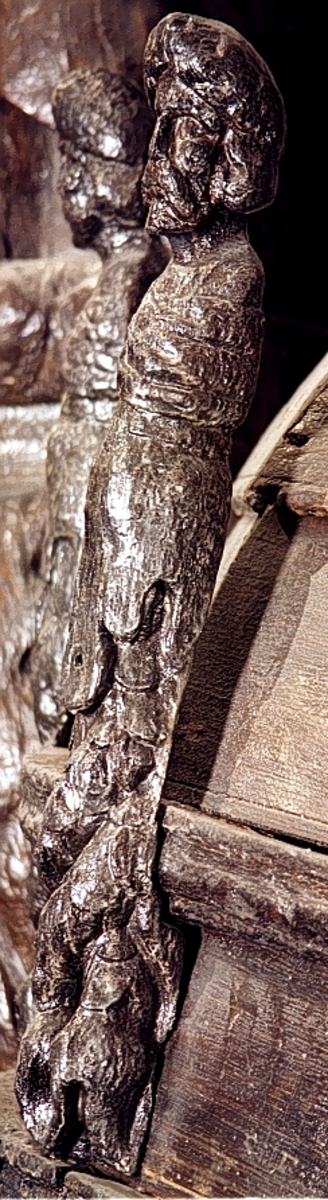 Skulpterad triton med turbanliknande lagerkrans på huvudet.

Huvudet har långt, ringlande hår samt stora och nedhängande mustascher. Snett över bröstet är en klädnad draperad. Klädnaden är fästad i en knut på höger axel. Kring livet sitter ett band med en oval.



Kroppens övergång till de dubbla, snodda fiskstjärtarna döljs av ett stort rundflikigt akantusblad. Huvudet är till största delen framställd som rundskulptur, dock något avplanad. Från nackhöjd och halvvägs ned på partiet med fiskstjärtarna är baksidan avplanad och slät - delvis något skålad - varefter det sitter ett snett urtag. Fotändan är vinkelformigt skålad vilket ger den en V-format tvärsektion, nedsänkningen är dock inte symmetriskt placerad. Nedtill böjer fotänden av inåt.

Skulpturen är mycket välbevarad.



Text in English: Sculpted Triton with a turban-like laurel wreath on its head.

The figure has a large pendulous moustache and long curly hair. There is a garment draped diagonally across the chest and fastened in a knot on the right shoulder. There is a girdle or belt round the waist with an oval centrepiece.



The transition between the body and the double fish tails is concealed by a large rounded acanthus leaf. The majority of the head is carved as a three-dimensional sculpture. The rear side surface has been planed smoothly.  The foot end has been hollowed out at an angle, giving it a V-shaped cross section, the hollowed-out section is not, however, symmetrically placed. The foot end curves inwards.

The sculpture is very well preserved.