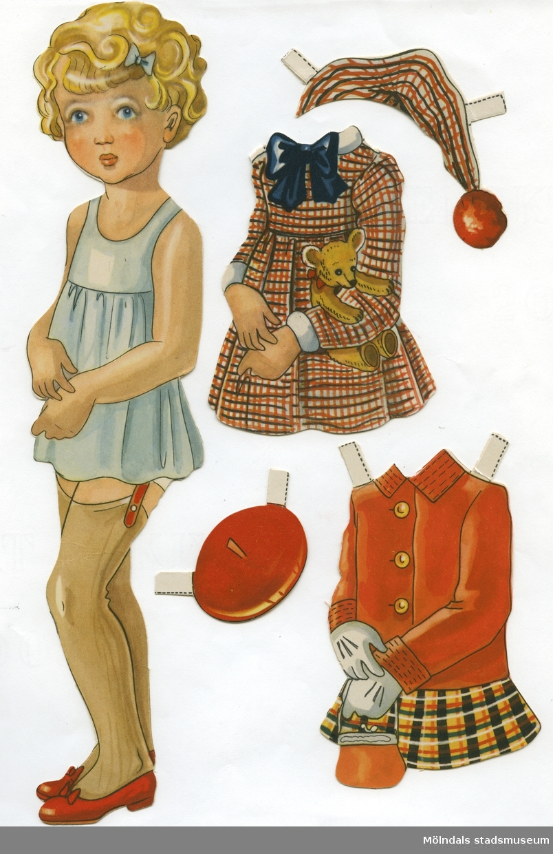Pappdocka med kläder från 1950-talet. Docka och kläder är märkta "Karin" på baksidan - dockans namn. Dockan föreställer en flicka med blond frisyr och blå ögon, iklädd underklänning, nylonstrumpor med strumpeband och skor. Garderoben består av tre klänningar, varav en med matchande mössa, kappa och kjol med basker, samt vinterkappa med byxor, pampuscher (galosch med pälsbräm), muff och hatt. Docka och kläder förvaras i en avlång påse av smörpapper, med texten "Påklädningsdockan 'Karin'".