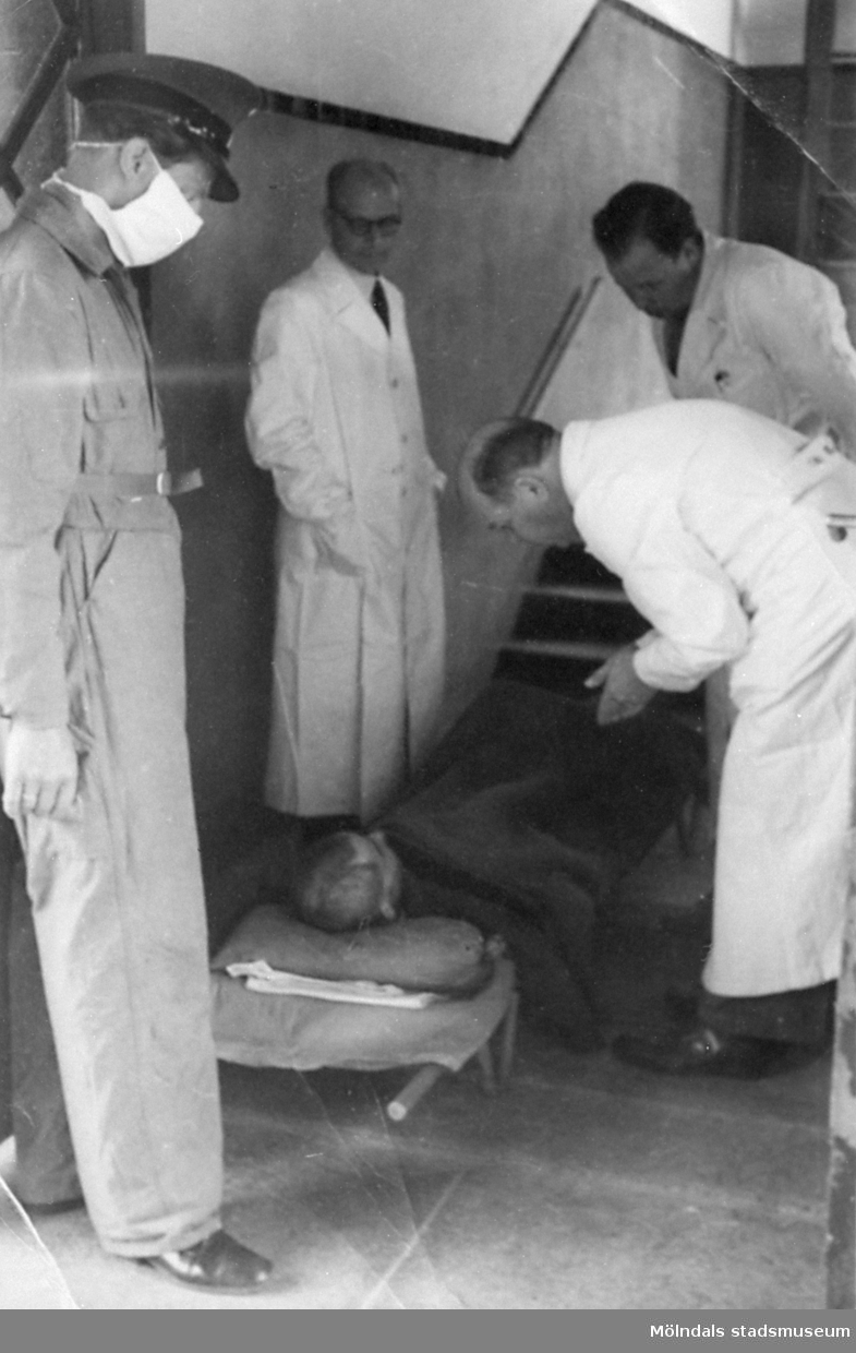 "Karantänsförläggning" på Kvarnbyskolan för överlevande från koncentrationsläger i Tyskland och Polen 1945.  
Skolan fungerade under denna sommar som beredsskapssjukhus.

En överlevande ligger på en bår, omgiven av tre läkare och en Röda Kors-arbetare.