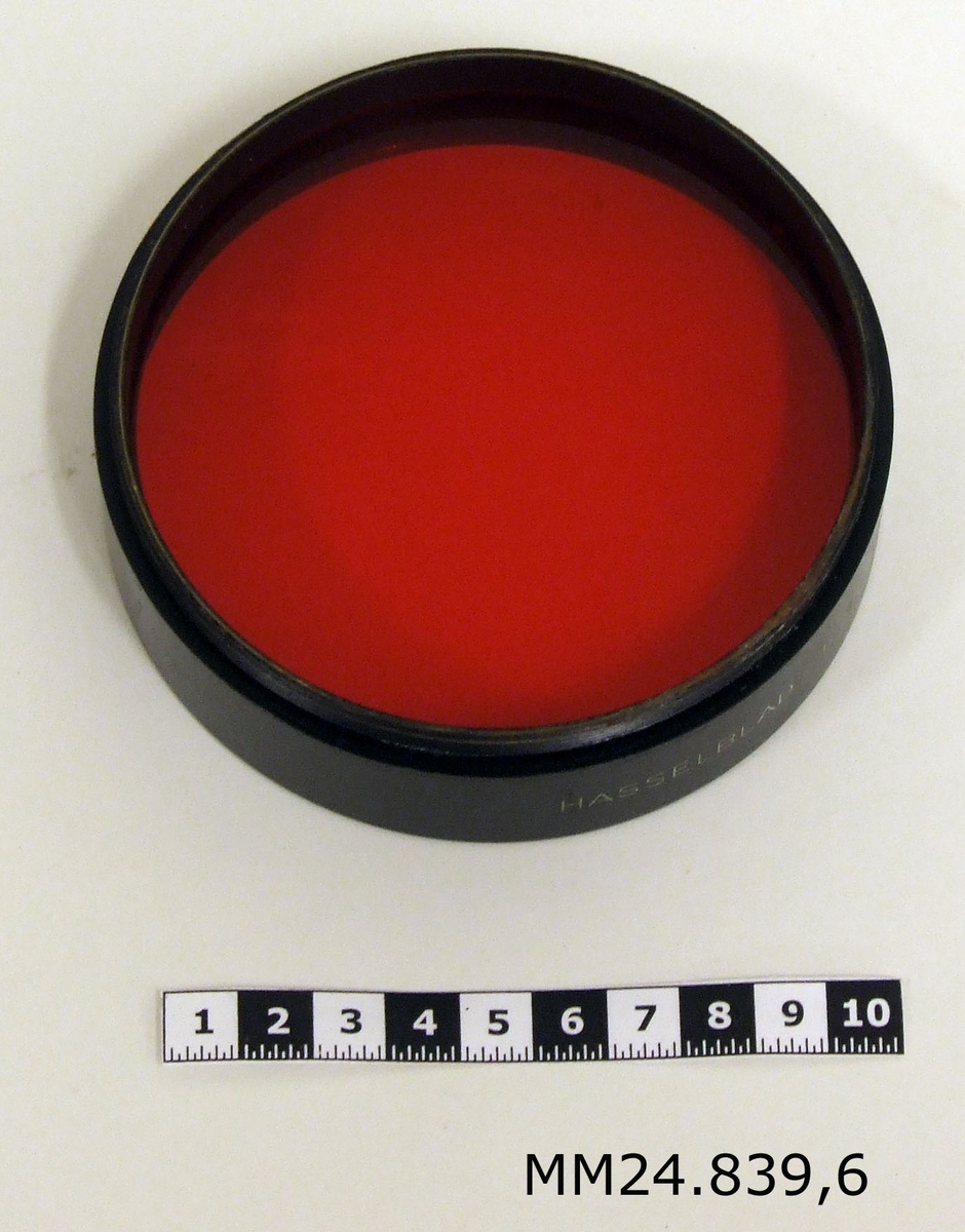 Cylinderformat filter av svart metall med rött glas. Märkning: "HASSELBLAD-R6-100 Made in Germany."